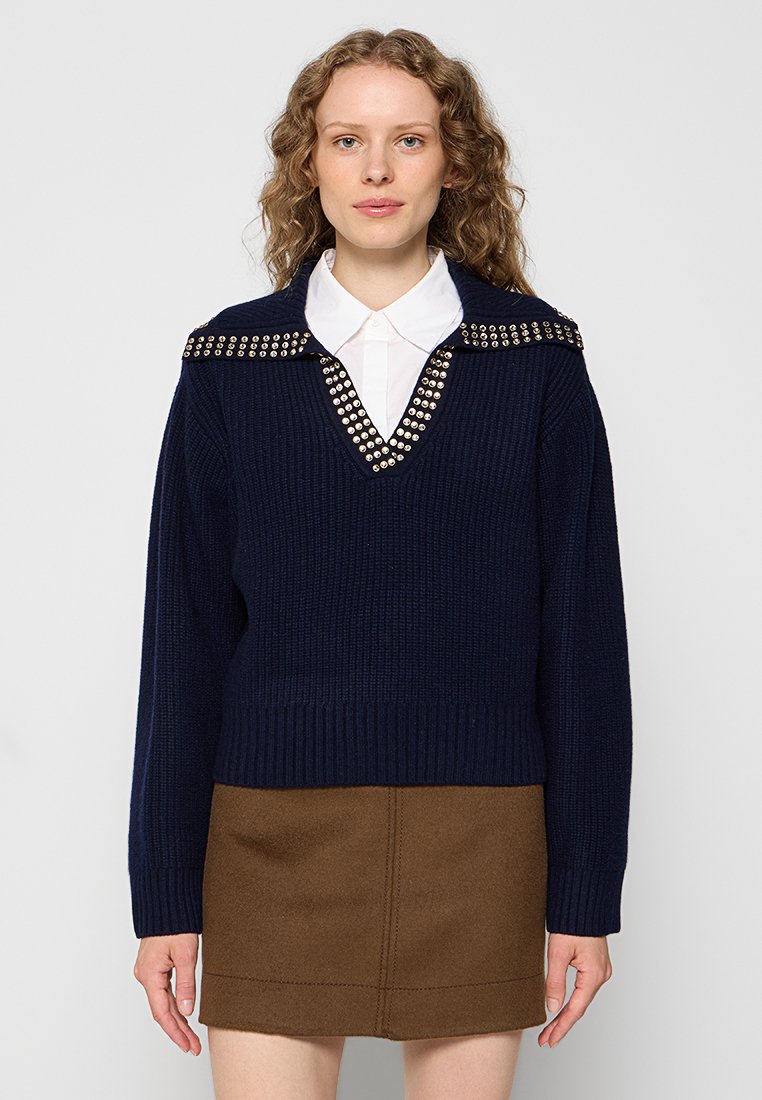 Pull en maille bleu marine avec un col en V et des accents d'épaules cloutés, associé à une jupe marron. Texture côtelée et coupe ajustée.