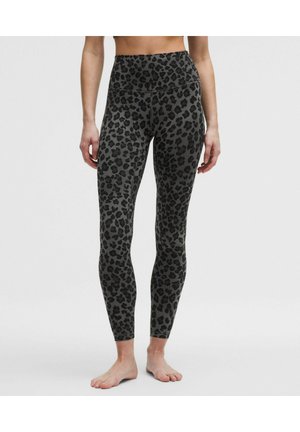 Frau trägt hochtaillierte, bodenlange Leggings mit schwarzem und grauem Leopardenmuster, steht barfuß vor einem schlichten Hintergrund.