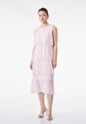 Frau in ärmellosem, hellrosa, gestuftem Midikleid und weißen High-Heel-Sandalen, mit neutralem Gesichtsausdruck vor weißem Hintergrund stehend.