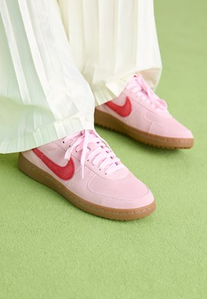 Baskets en daim rose avec un logo rouge et des lacets assortis. La semelle est en caoutchouc brun clair. Portées avec un pantalon plissé blanc sur une surface verte.