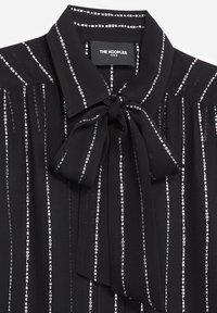 Blusa negra con cuello y detalle de lazo, presenta patrones de texto vertical en blanco en toda su superficie. Fabricada en una tela suave con una textura lisa.