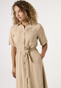 Beige Blusenkleid mit kurzen Ärmeln, frontalen Taschen und einem gebundenen Gürtel in der Taille. Aus weichem Stoff gefertigt und mit einer lockeren Passform.