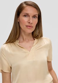 Blouse beige à manches courtes avec col à notch, texture douce et éclat subtil ; fabriquée dans un tissu léger avec des coutures minimales.