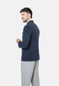 Blazer azul marino con diseño estructurado, cierre de un solo botón y bolsillo en el pecho. Combinado con pantalones gris claro, que destacan un corte a medida.