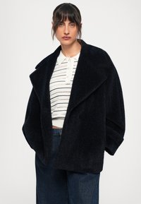 Casaco oversized em mistura de lã azul-marinho, com colarinho notched, tecido texturado e grandes lapelas. Usado sobre uma camisola branca com riscas e jeans.