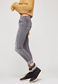 Jeans skinny grises con detalles desgastados, combinados con zapatillas negras que tienen una textura de punto y una banda blanca en el tobillo.