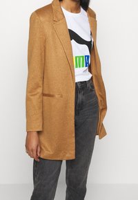 Lång, skräddarsydd blazer i ljusbrunt tyg med en slät textur och hakkragar. Kombineras med en vit t-shirt med grafik och mörka jeans.