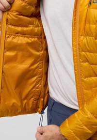 Veste en duvet jaune avec texture matelassée, fermeture éclair complète et ourlet ajustable. Portée sur une chemise de couleur claire, mettant en valeur la doublure intérieure.