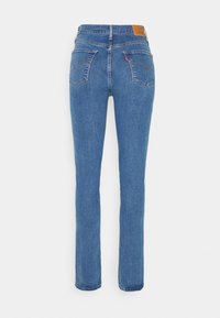 Jean denim bleu à jambes droites avec poches arrière, coutures orange, patch en cuir marron et étiquette rouge sur la poche droite sur fond blanc.