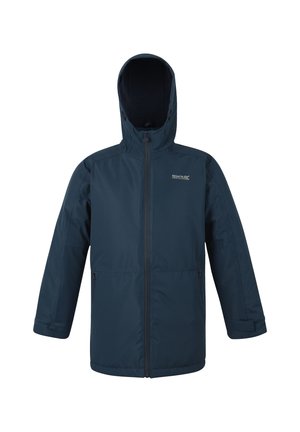 Młody chłopiec uśmiechający się, ubrany w niebieski płaszcz, białą koszulę, paskowany krawat, ciemne spodnie i czarne buty, trzymający niebieską butelkę na wodę.