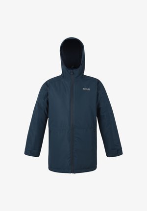 Młody chłopiec uśmiechający się, ubrany w niebieski płaszcz, białą koszulę, paskowany krawat, ciemne spodnie i czarne buty, trzymający niebieską butelkę na wodę.