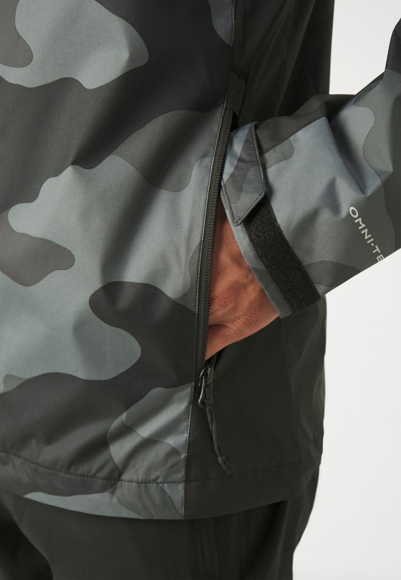 Chaqueta resistente al agua con un patrón de camuflaje gris oscuro, que cuenta con un bolsillo con cremallera y cierre de velcro en los puños. El material parece suave.