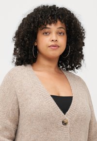 Béžový pletený cardigan s hlubokým výstřihem do V a jedním knoflíkem na zapínání. Měkká textura a žebrované okraje. Model má pod ním černý top.