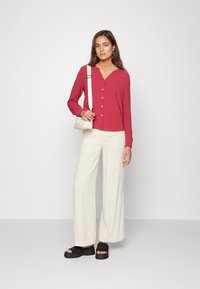 Camisa vermelha de manga longa com botões e decote em V, combinada com calças largas em creme e sapatos pretos sem fecho. Pequena bolsa de ombro usada a tiracolo.