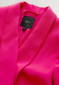 Růžový blejzr z hladké látky s klopovým límcem. Viditelná značka s nápisem "NEXT." Čistý, moderní design bez ozdob.