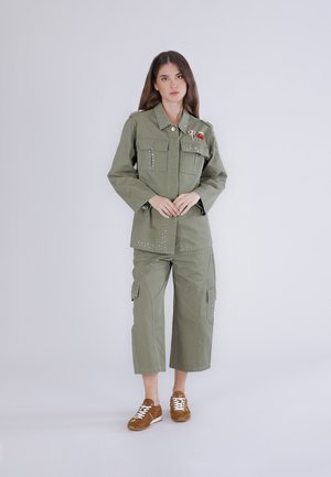 Kvinde stående iført olivengrøn nittebesat arbejdstøjjakke og korte cargobukser med brune sneakers mod en ensfarvet baggrund.