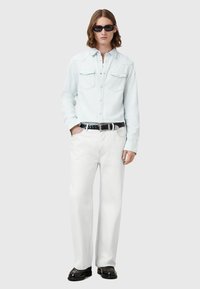 Lysblå skjorte med knapper og to brystlommer, hvide vidde jeans, sort fletbælte og sorte sko. Model iført mørke solbriller.