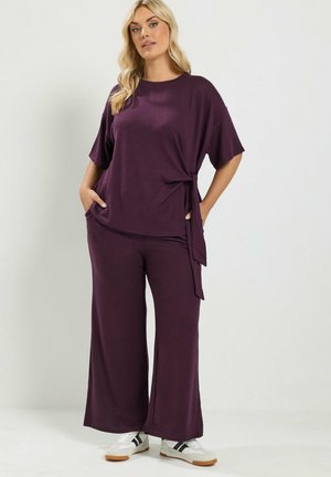 Femme aux longs cheveux blonds porte un haut ample violet foncé et un pantalon large assorti avec des baskets blanches, debout devant un fond clair uni.
