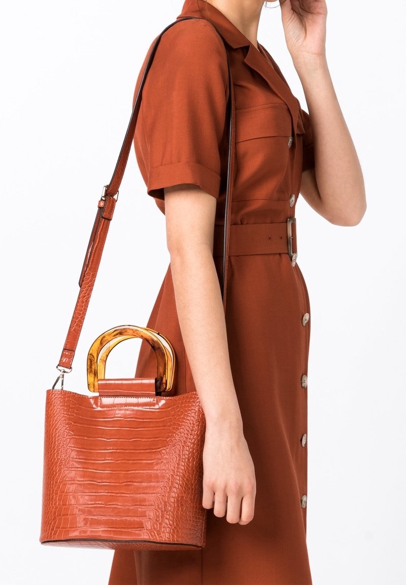 Femme en robe rouille à manches courtes tenant un sac à main marron texturé avec une épaisse poignée effet marbre et une bandoulière, debout sur un fond blanc.