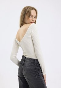Long-sleeve, off-white ribbed top with a deep V-back, paired with high-waisted, dark gray jeans. Smooth fabric and fitted design.