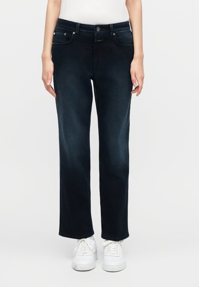 Femme portant un jean droit bleu foncé et des baskets blanches, debout devant un fond clair uni.