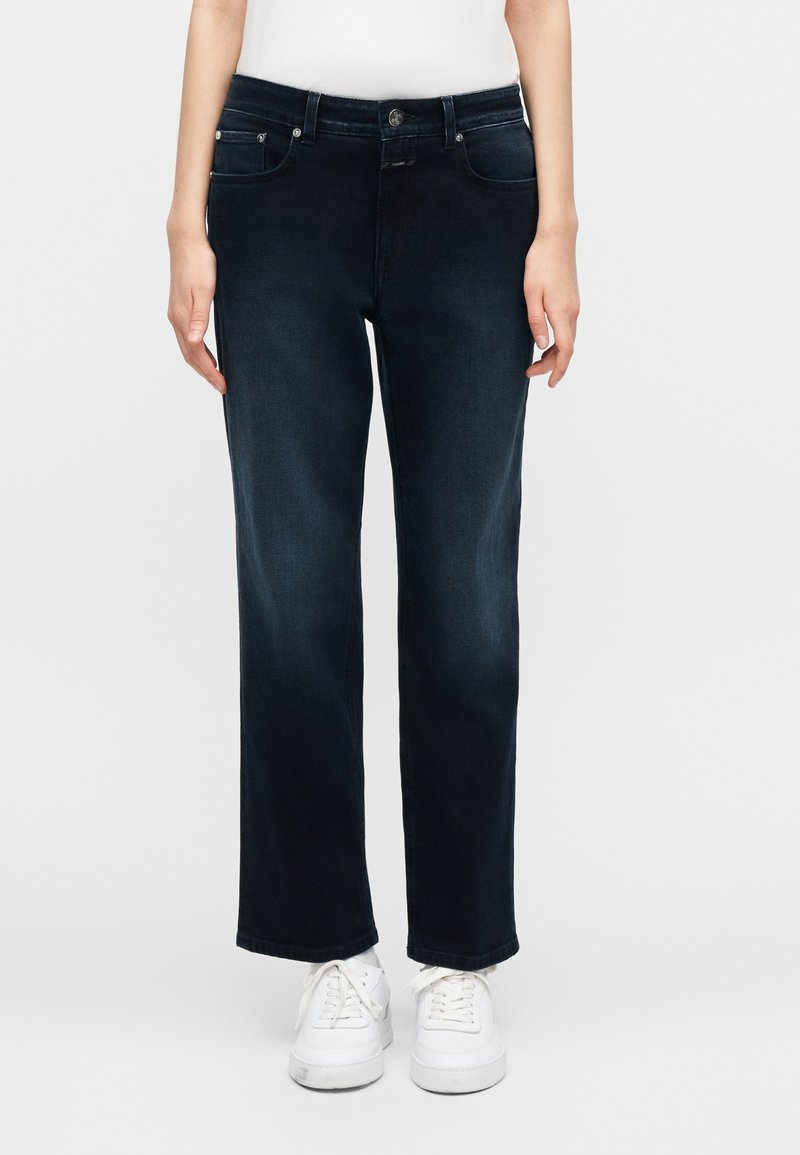 Femme portant un jean droit bleu foncé et des baskets blanches, debout devant un fond clair uni.