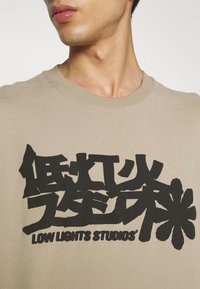 Gros plan d'une personne portant une chemise beige avec de grands caractères asiatiques noirs stylisés et le texte "LOW LIGHTS STUDIOS" à l'avant.
