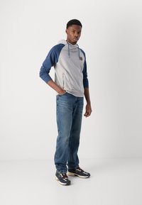 Sudadera gris y azul marino con un bolsillo frontal, cordones y detalle del logotipo, combinada con jeans azul claro y zapatillas multicolor.