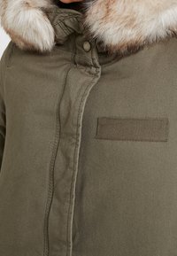 Olivgrüne Jacke mit einem Kragen aus Kunstfell, ausgestattet mit einem Knopfverschluss und einem kleinen rechteckigen Aufnäher auf der linken Seite. Glatte, matte Textur.
