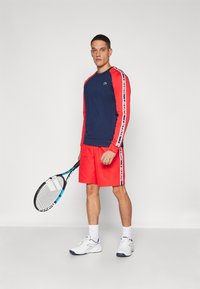 Marinblå langermet topp med røde ermer, med logodetalj; røde shorts med sidestriper. Holder en tennissracket, iført hvite joggesko.