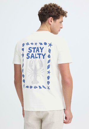 Mann trägt ein weißes T-Shirt mit einer blauen Hummer-Grafik und Muschelrand, Rückseite mit dem Schriftzug "STAY SALTY", zur Seite blickend.