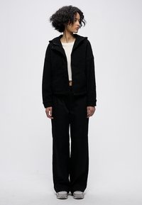 Melnas vilnas jaka ar kapuci, rāvējslēdzēju un sānu kabatām, kombinēta ar plaša piegriezuma melnām biksēm un pelēkām kurpēm. Minimālistisks dizains.