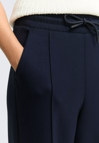 Hosen in Marineblau aus glattem Stoff mit einem elastischen Bund und Kordelzug, seitlichen Taschen und zusätzlichen Falten an der Vorderseite für mehr Detail.