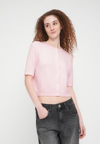 Γυναίκα με σγουρά κόκκινα μαλλιά που φοράει ένα ανοιχτό ροζ κοντομάνικο ζακέτα με κουμπιά και μαύρο ξεβαμμένο τζιν με το ένα χέρι στην τσέπη.