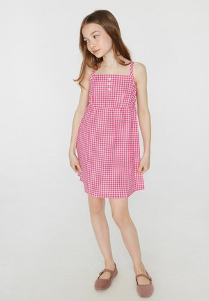 Jeune fille aux longs cheveux bruns portant une robe sans manches à carreaux rose et des chaussures Mary Jane marron, debout sur un fond blanc uni.