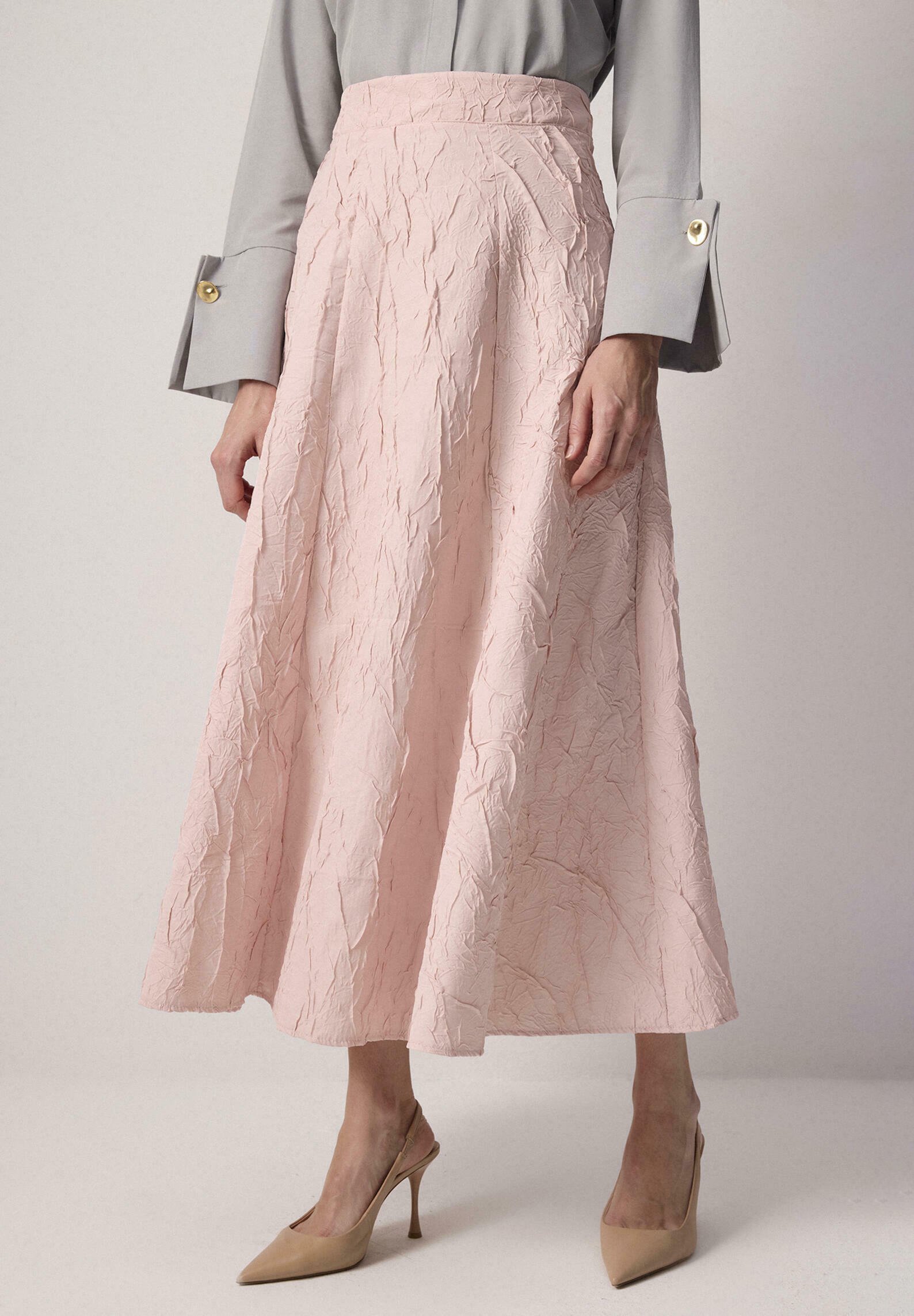 Femeie purtând o fustă midi roz pal cu cute, o bluză gri cu manșete cu nasturi aurii și pantofi slingback bej cu vârf ascuțit, stând pe un fundal simplu.