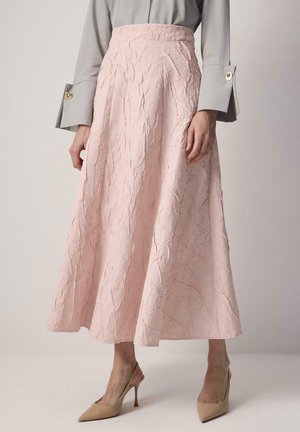 Ženska, oblečena v nagubano bledo rožnato midi krilo, sivo bluzo z zlatimi gumbi na manšetah in gole zarobljene pete z odprtim petnim trakom, stoji na enostavni podlagi.