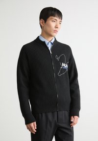 Svart strikket cardigan med glidelås foran, ribbestruktur og brodert design. Har høy krage og lange ermer. Kombinert med en blå skjorte.