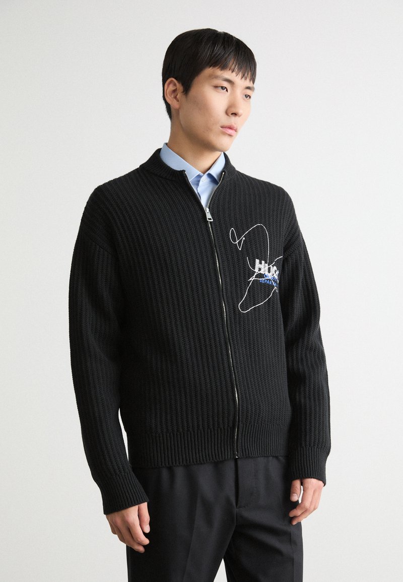 Svart strikket cardigan med glidelås foran, ribbestruktur og brodert design. Har høy krage og lange ermer. Kombinert med en blå skjorte.