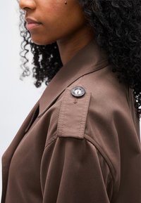 Gros plan d'une personne aux cheveux bouclés portant une veste marron avec un détail de patte d'épaule boutonnée.