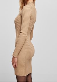 Abito bodycon in maglia color beige, con texture a costine, maniche lunghe, colletto alto e silhouette aderente con orlo scallop.