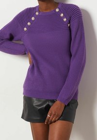 Paarse gebreide trui met ribbelstructuur, voorzien van knoopaccenten op de schouders en lange mouwen. Gecombineerd met een zwarte leren minirok.