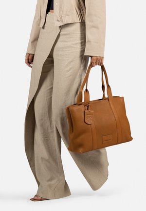 Sac tote en cuir brun avec une forme structurée, des poignées doubles et un logo embossé. Accompagné d'un pantalon à jambes larges de couleur claire.