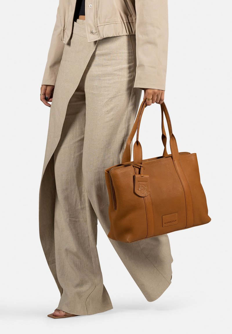 Sac tote en cuir brun avec une forme structurée, des poignées doubles et un logo embossé. Accompagné d'un pantalon à jambes larges de couleur claire.