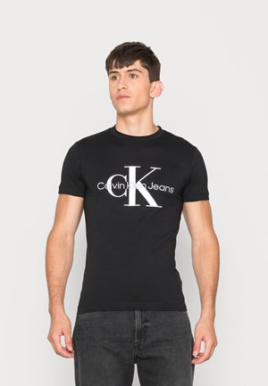 Mladi muškarac nosi usku crnu Calvin Klein Jeans majicu s velikim bijelim "cK" logotipom, stoji ispred jednostavne svijetle pozadine.