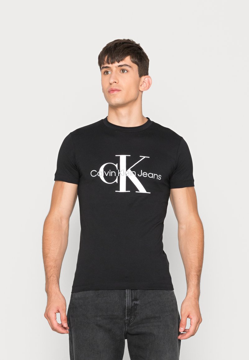 Young man wearing a fitted black Calvin Klein Jeans t-shirt with large white "cK" logo, standing against a plain light background.