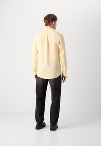Yellow linen shirt with long sleeves, a button-down collar, and a curved hem, paired with black trousers and black shoes.