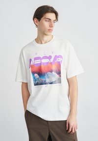 Fehér pamut póló, színes grafikával, amely felhőket és lila "FLY ABOVE" feliratot ábrázol. Rövid ujjú, laza szabású.