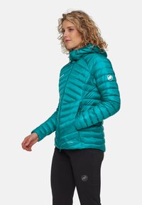 Kvinna med lockigt hår som bär en teal-färgad quiltad jacka med huva och svarta byxor, står sidan mot med ena handen i fickan.