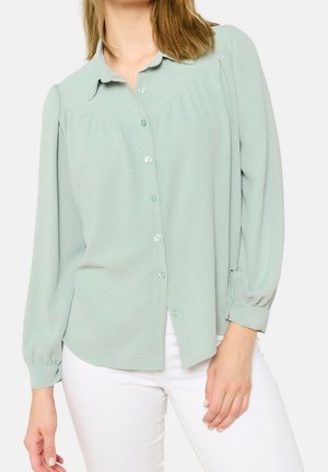 Frau trägt eine hellgrüne, langärmelige Bluse mit Knöpfen und weiße Hose, steht vor einem schlichten Hintergrund.